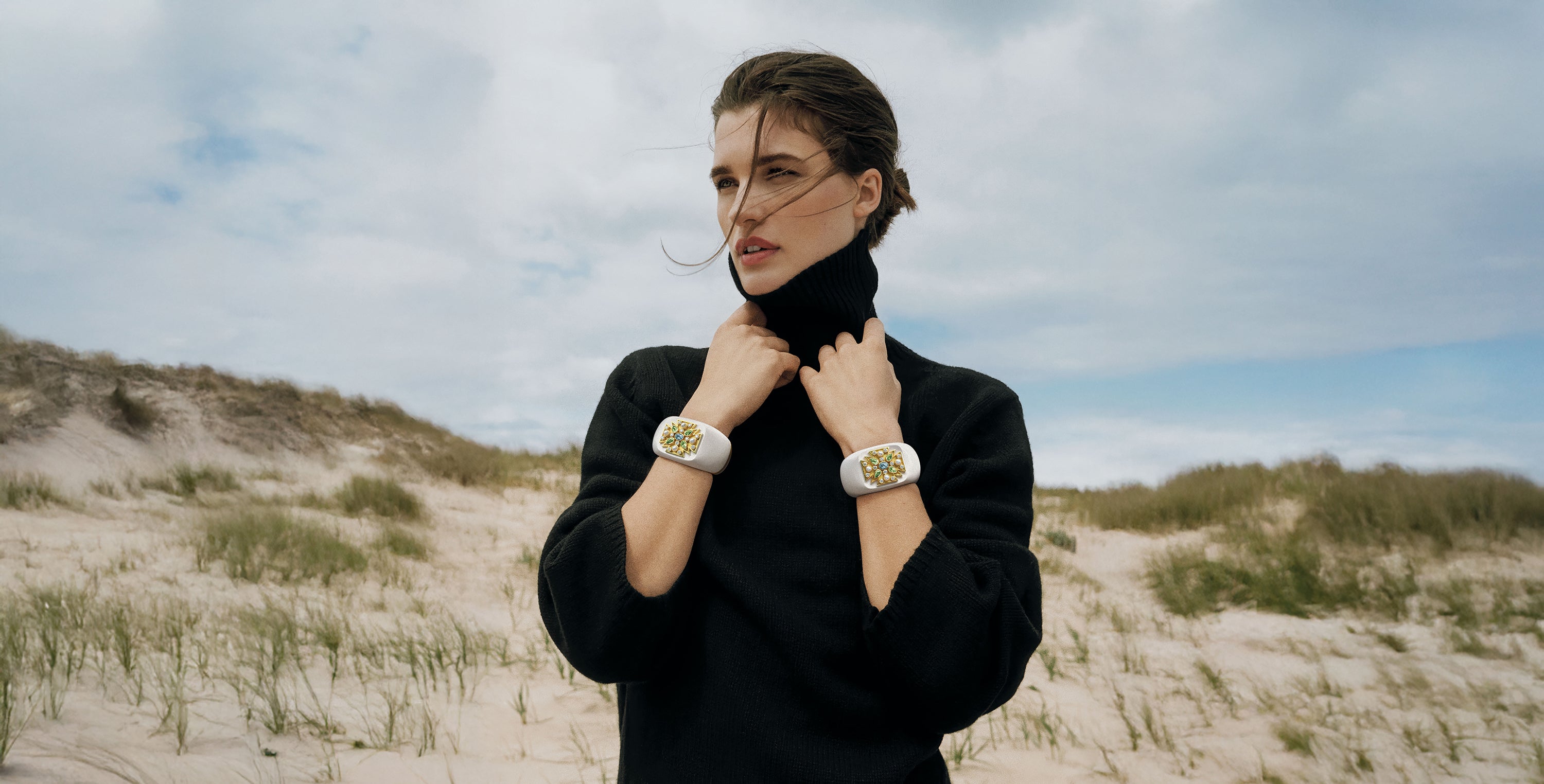 Woman in black outfit standing on a sandy dune with a cloudy sky wearing two Verdura Maltese Cross Cuffs