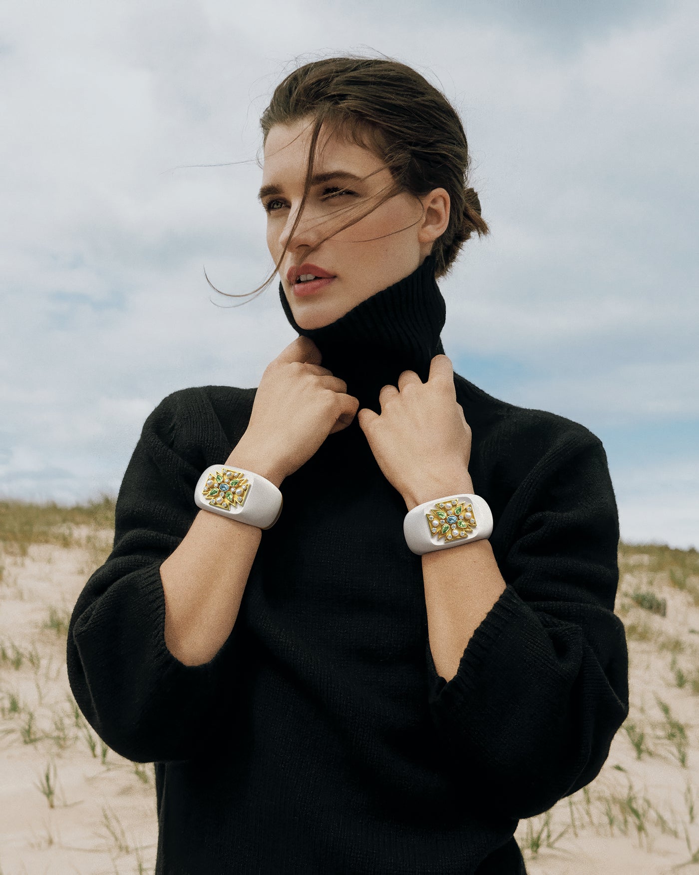 Woman in black outfit standing on a sandy dune with a cloudy sky wearing two Verdura Maltese Cross Cuffs